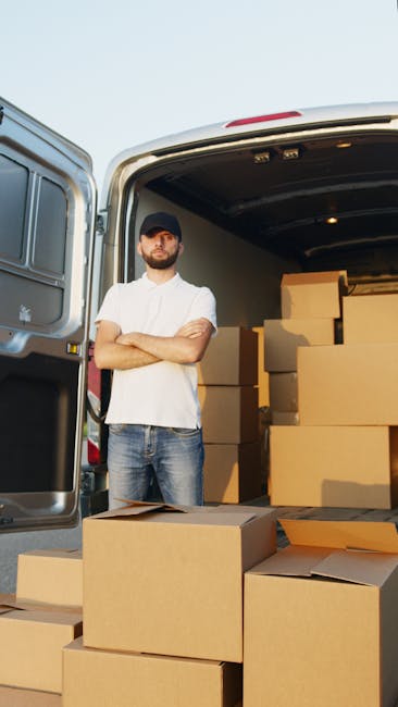 A man with a beard, wearing a dark cap, white t-shirt, and jeans, standing with arms crossed next to the open rear doors of a white moving van. The van's interior is visible, showing multiple stacked 