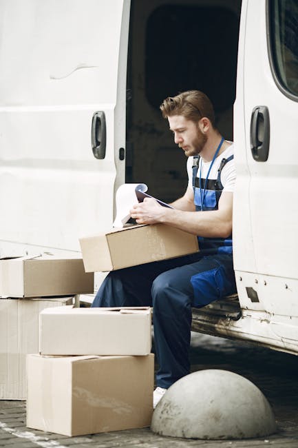 A man with a beard, wearing a dark cap, white t-shirt, and jeans, standing with arms crossed next to the open rear doors of a white moving van. The van's interior is visible, showing multiple stacked 
