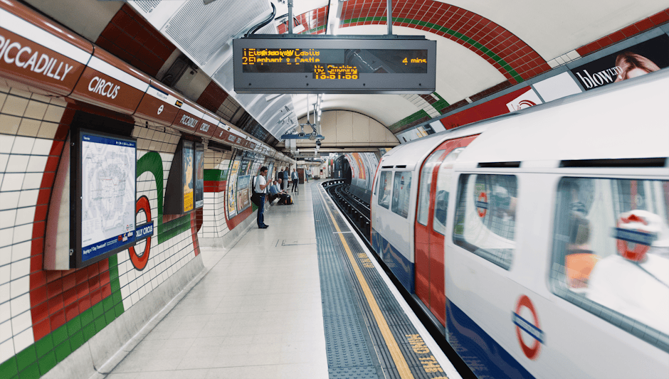 Inside the Colliers Wood underground station platform with a moving train arriving on the right, featuring the station's red, white, and blue roundel logo on the train's exterior. The platform has white tiled walls with green and red accents, and an electronic departure board displaying train information and timing. Several passengers are standing or sitting along the platform, while some are engaged in packing or preparing for home relocation, with luggage and shopping bags visible. The platform floor is grey, with tactile paving near the edge for safety, and the station environment is well-lit with overhead lighting. The scene captures the typical setting for people traveling to or from a house removals or furniture transport service near Colliers Wood Tube Station, as provided by [COMPANY_NAME].