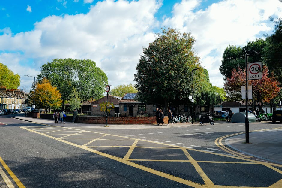 A street scene near Colliers Wood Tube Station featuring a small brick building surrounded by tall, leafy trees with green and autumnal foliage. Several pedestrians are walking on the sidewalk, some accompanied by bicycles or scooters parked nearby. The road is marked with yellow box junction lines and a 20 mph zone sign is visible on a lamp post. The sky overhead is partly cloudy with patches of blue, and there are parked cars along the street. The environment suggests a quiet residential or local area, where house removals and furniture transport activities are often coordinated by companies specializing in home relocation services.