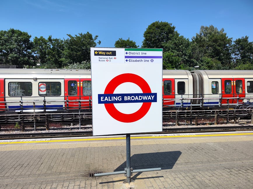 Inside the Colliers Wood underground station platform with a moving train arriving on the right, featuring the station's red, white, and blue roundel logo on the train's exterior. The platform has white tiled walls with green and red accents, and an electronic departure board displaying train information and timing. Several passengers are standing or sitting along the platform, while some are engaged in packing or preparing for home relocation, with luggage and shopping bags visible. The platform floor is grey, with tactile paving near the edge for safety, and the station environment is well-lit with overhead lighting. The scene captures the typical setting for people traveling to or from a house removals or furniture transport service near Colliers Wood Tube Station, as provided by [COMPANY_NAME].