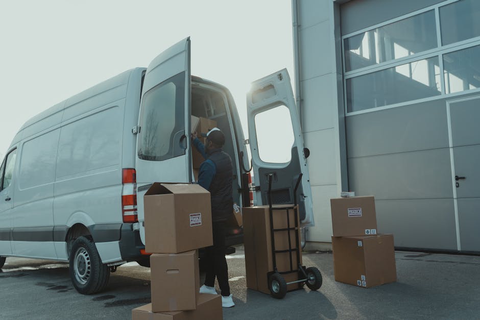 A professional mover from [COMPANY_NAME] is engaged in a home relocation process, standing beside a white delivery van parked outside a modern building with large glass windows. The mover, wearing a dark uniform with a cap, is loading or unloading cardboard boxes into the back of the van, which has its rear doors open to reveal interior packing materials. Several additional cardboard boxes, some sealed with packing tape and labeled, are placed on the pavement nearby. A wheeled trolley or dolly is positioned next to the boxes, ready for transport. The scene takes place during daylight with clear visibility and some sunlight creating glare behind the van. The environment suggests an organized loading process, with early stages of furniture transport and packing activities associated with a house removal service, supported by [COMPANY_NAME], highlighting efficient logistics and safe handling of belongings for a move on Colliers Wood High Street.
