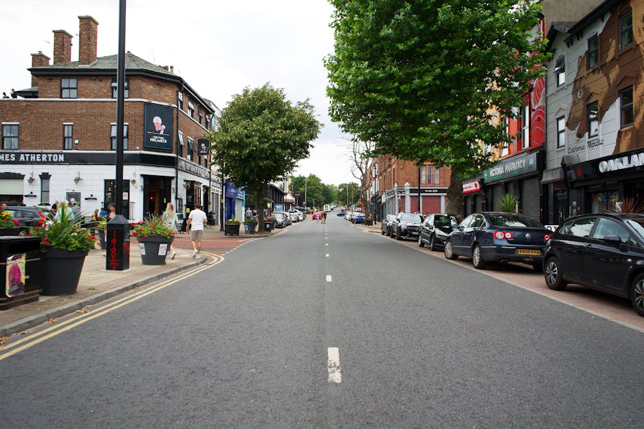 A wide street scene on Colliers Wood High Street, featuring parked cars lining both sides of the road, with a variety of shops and businesses such as a pharmacy, cafes, and retail outlets visible on the buildings. The road has a single dashed white line in the centre, and the pavement on each side is occupied by pedestrians, some walking and others standing or sitting near large black planters filled with colorful flowers. Several trees with full green foliage are positioned along the sidewalk, providing shade. The street appears calm and busy, with a clear sky overhead. This setting provides a backdrop for local house removals or furniture transport, with an emphasis on urban logistics and loading procedures typical of a home relocation or moving service on Colliers Wood High Street, as managed by [COMPANY_NAME].