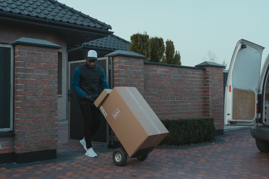 A professional mover from [COMPANY_NAME] is engaged in a home relocation process, standing beside a white delivery van parked outside a modern building with large glass windows. The mover, wearing a dark uniform with a cap, is loading or unloading cardboard boxes into the back of the van, which has its rear doors open to reveal interior packing materials. Several additional cardboard boxes, some sealed with packing tape and labeled, are placed on the pavement nearby. A wheeled trolley or dolly is positioned next to the boxes, ready for transport. The scene takes place during daylight with clear visibility and some sunlight creating glare behind the van. The environment suggests an organized loading process, with early stages of furniture transport and packing activities associated with a house removal service, supported by [COMPANY_NAME], highlighting efficient logistics and safe handling of belongings for a move on Colliers Wood High Street.