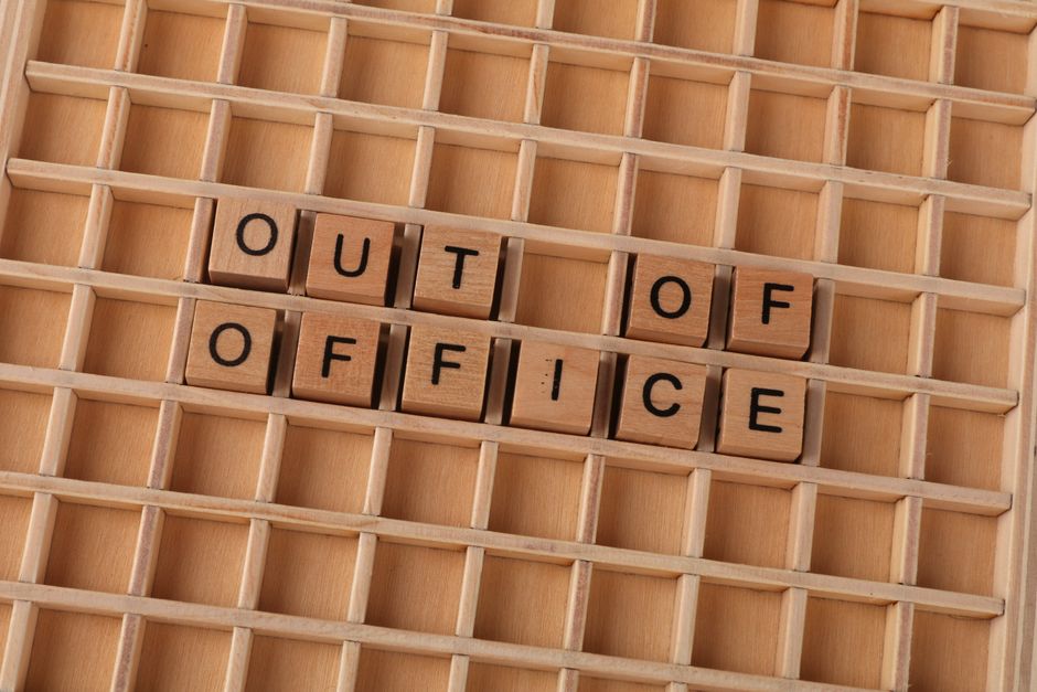 A close-up view of a wooden grid display board with small square compartments. Sixteen compartments are visible, with the middle row spelling out the phrase 'OUT OF OFFICE' using small wooden letter blocks with black letters on a light wood surface. The background shows a plain, light wood finish, and the overall scene suggests organization or labeling, often associated with office or workplace environments. The image is taken indoors, with even lighting highlighting the natural wood textures. This visual relates to the concept of office removals or business relocations, where clear labeling and packing are essential, and is provided as part of professional house or office moving services, such as those by removal companies like removalcompaniescollierswood.co.uk.