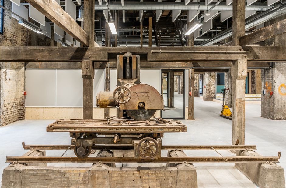 Inside an industrial building with exposed brick walls and steel ceiling beams, there is an old, rusted circular saw machine mounted on a wooden platform with metal wheels and rails. The machine is positioned on a concrete floor, with various wooden and metal components nearby. In the background, there are open doorways leading to other sections of the building, and a modern yellow floor scrubber is visible on the right side, indicating cleaning or maintenance activities. The space is well-lit, with natural and artificial lighting illuminating the area, and there are wooden and metal structures supporting the upper level or mezzanine. The scene appears to be an early stage of building renovation or industrial clearance, with no furniture or boxes present for house or office removals, but the photograph exemplifies the working environment involved in moving or demolishing industrial equipment or preparing premises for a house or business relocation, as handled by [COMPANY_NAME].